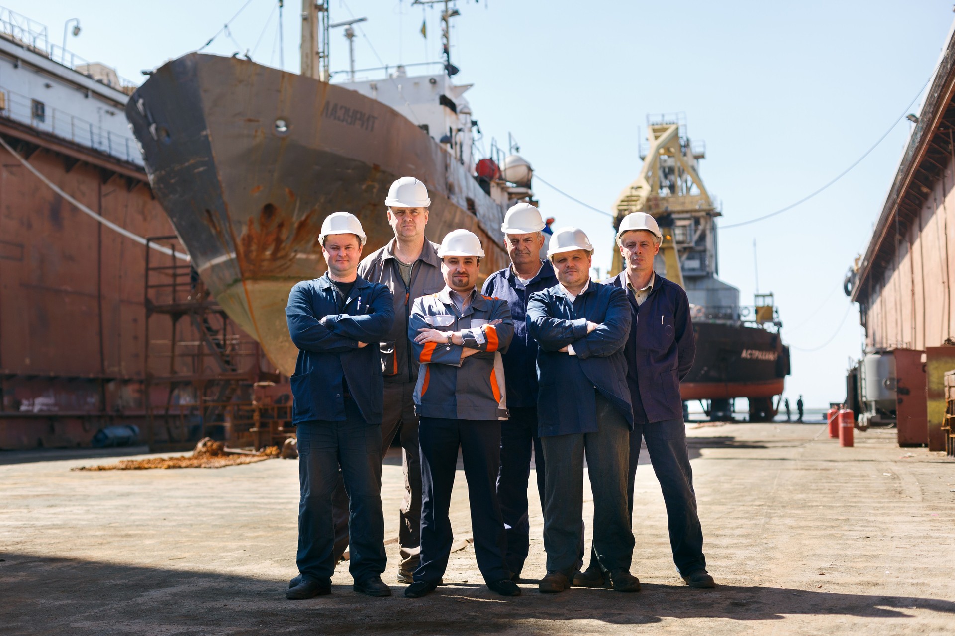 Team of shipyard workers stand proudly at dry dock. Marine engineers in helmets, repair crew with vessel. Dockyard laborers by ship hull, maritime industry, maintenance on sunshine day.